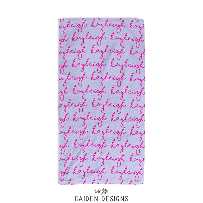 A personalized beach towel featuring a repeating pattern of the name 'Kayleigh' in bright pink cursive script on a light lavender background. The brand name 'CAIDEN DESIGNS' is printed below the product on a white background.