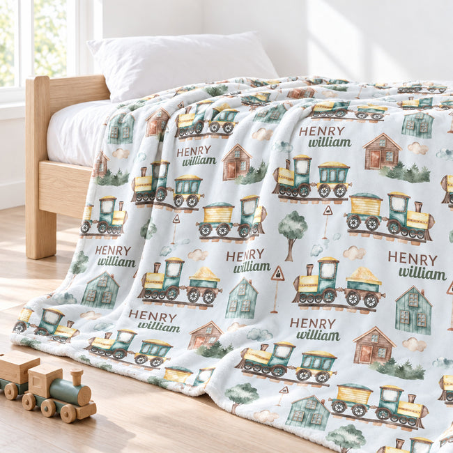 A personalized blanket with a train and house pattern, draped over a wooden bed in a bright, sunlit bedroom. The blanket features the name 'HENRY william' printed repeatedly alongside illustrations of colorful toy trains, small houses, and trees.