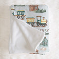 A folded baby blanket with a white sherpa backing and a printed cotton front featuring vintage watercolor trains, houses, and text, displayed on a white faux fur background.