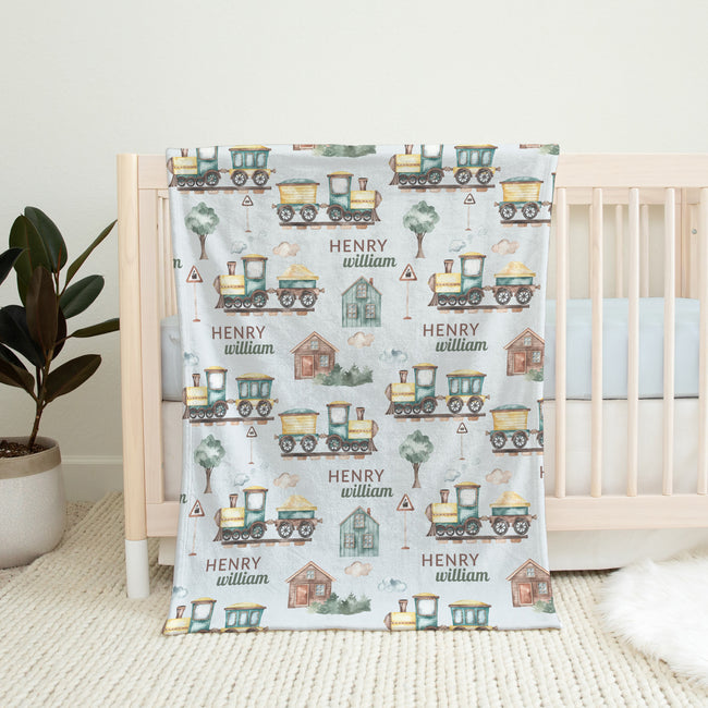 A personalized baby blanket with a vintage train pattern and the name 'HENRY william' printed on it, draped over a light wood crib in a nursery setting.