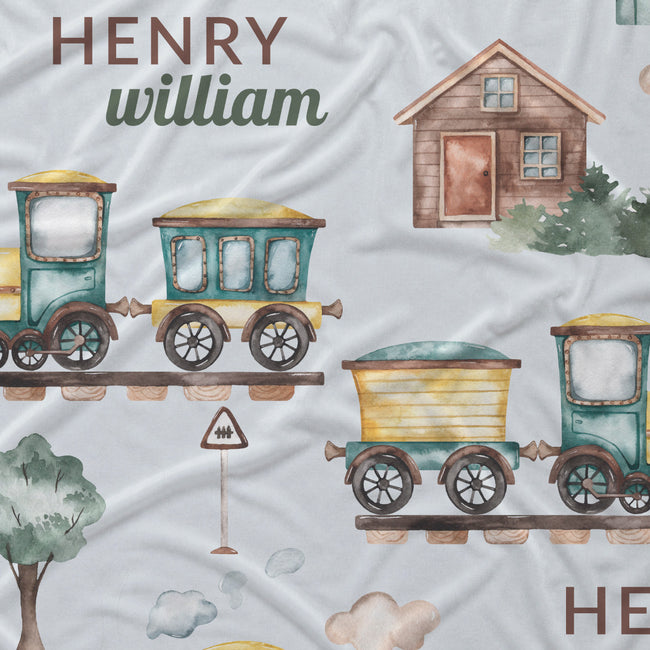 Personalized baby bedding fabric featuring a watercolor train pattern with the name 'HENRY' and 'william' in brown and green lettering, alongside a small house and trees on a light gray background.