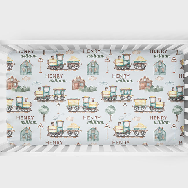 A crib sheet featuring a repeating pattern of vintage toy trains, small houses, trees, and the names 'HENRY' and 'william' on a pale blue background, shown inside a white crib.