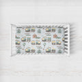 A white wooden crib with a fitted sheet featuring a repeating pattern of vintage-style trains, houses, trees, and the names 'HENRY' and 'william' on a light gray background.