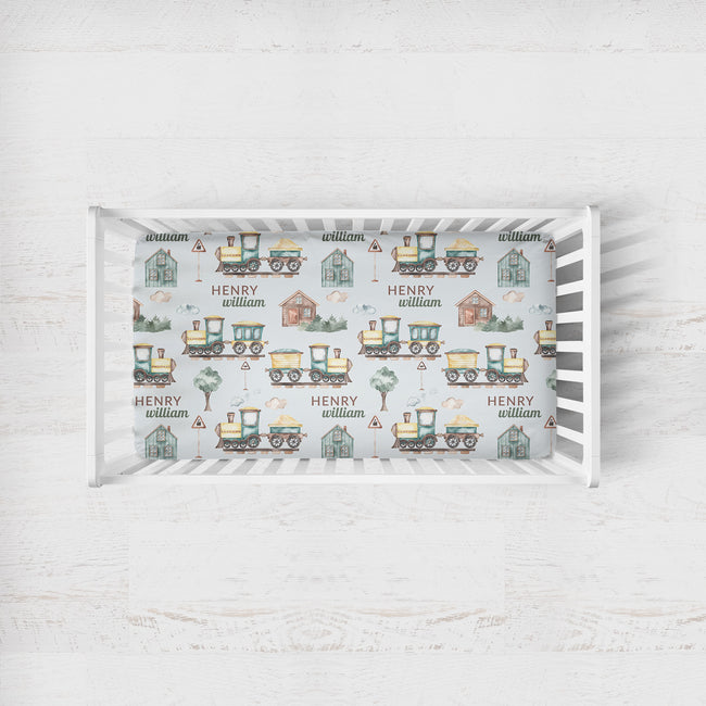 A white wooden crib with a fitted sheet featuring a repeating pattern of vintage-style trains, houses, trees, and the names 'HENRY' and 'william' on a light gray background.