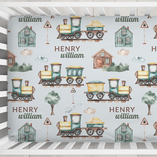 A personalized baby crib mattress cover featuring a repeating pattern of vintage toy trains, small houses, trees, and the names 'HENRY' and 'william' printed in brown text on a pale blue background.