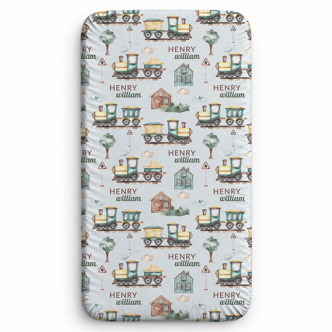 A personalized fitted sheet with a repeating pattern of vintage yellow trains, a small red barn, trees, and the names 'HENRY' and 'william' on a light gray background.