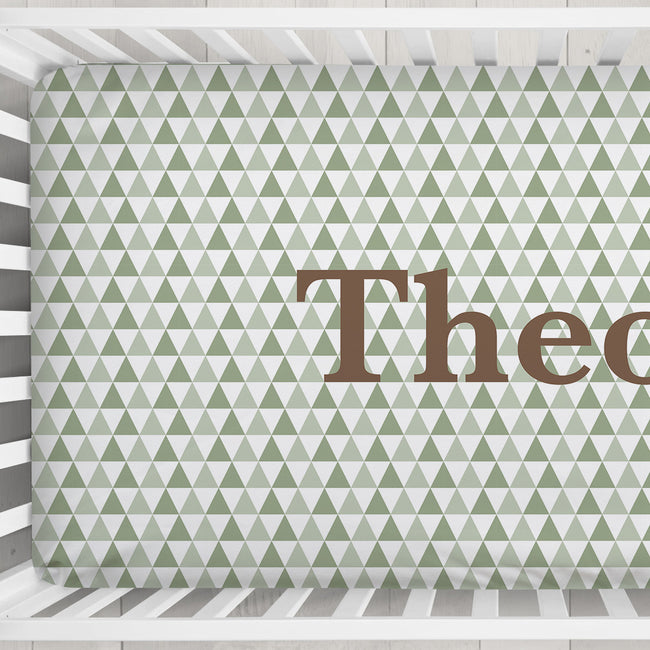 A crib sheet featuring a repeating pattern of small green and white triangles with the partial brown text 'Theo' printed across it, shown inside a white wooden crib.