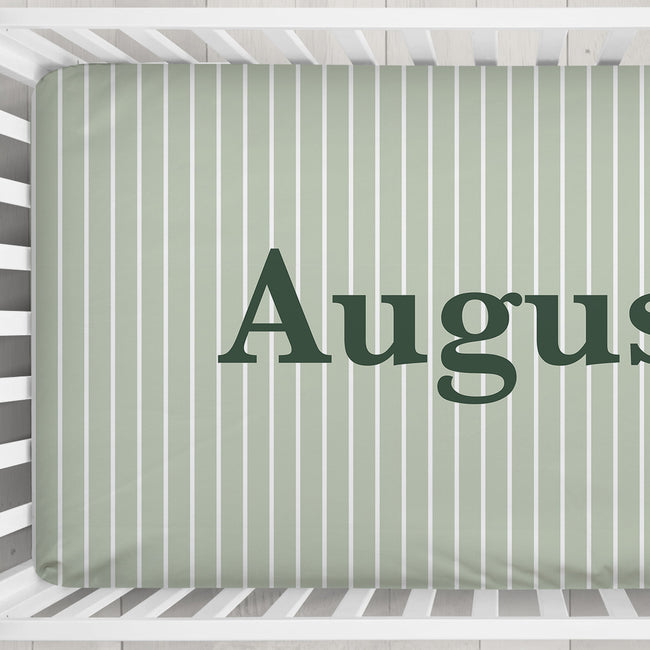 A close-up of a crib mattress cover with vertical white stripes on a sage green background, featuring the partial name 'August' in dark green serif lettering.