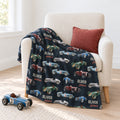 Personalized vintage race car blanket featuring the name Oliver James, draped over a cream kids’ chair with a rust pillow and blue toy race car.