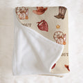A folded fleece blanket with a western-themed print on one side and a soft white sherpa lining on the reverse, displayed on a white faux fur background.