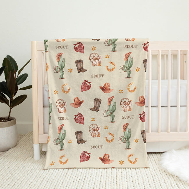 A cream-colored baby blanket with a rustic western-themed print featuring cowboy boots, cacti, horseshoes, and the word 'SCOUT', draped over a light wood crib in a nursery setting.