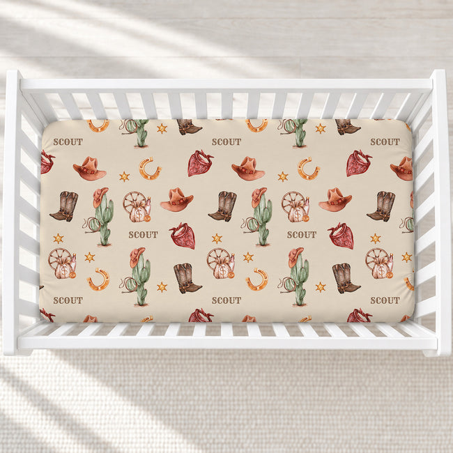 A fitted crib mattress with a western-themed Scout pattern, shown inside a white wooden crib. The pattern features cacti, cowboy hats, boots, horseshoes, and stars in earthy brown, orange, and green tones on a cream background, with the word 'SCOUT' printed repeatedly.