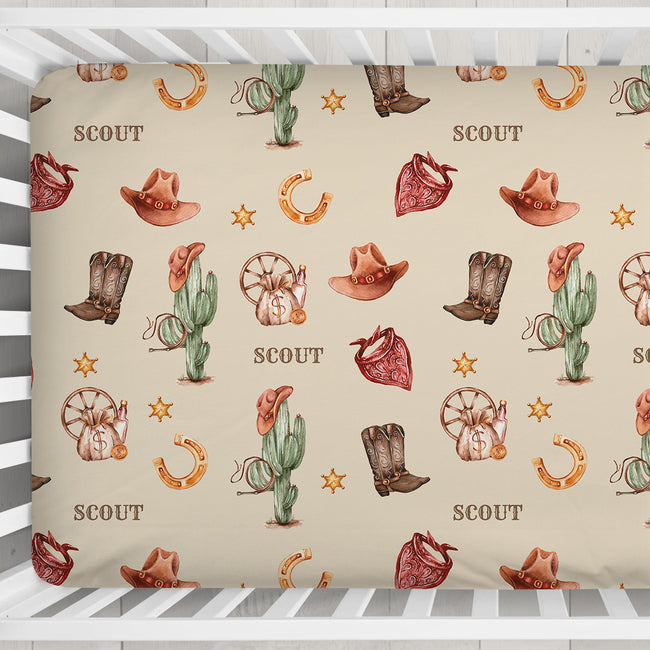 Overhead view of a Wild West crib sheet personalized with Scout, featuring cowboy boots, hats, cacti and bandanas inside a white crib.