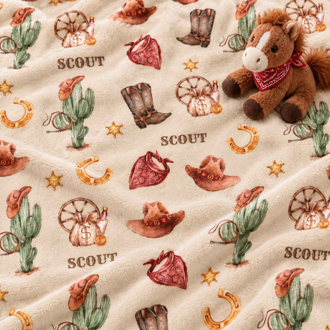 A plush fleece blanket with a western 'SCOUT' theme, featuring a brown plush toy horse on the right side. The blanket has a cream-colored background with a repeating pattern of brown cowboy hats, green cacti, red bandanas, brown boots, gold horseshoes, and brown wagon wheels, with the word 'SCOUT' printed in brown text.