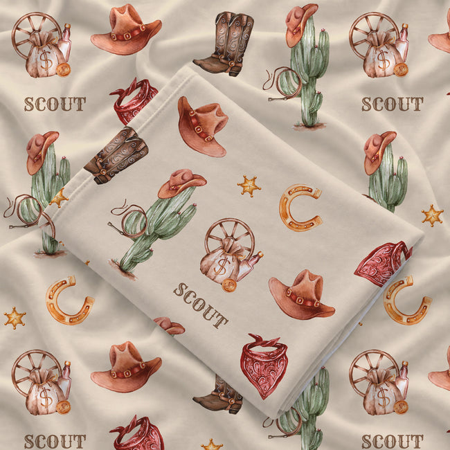 Folded Wild West swaddle blanket showing Scout personalization, cowboy illustrations and stitched edging on cream fabric.