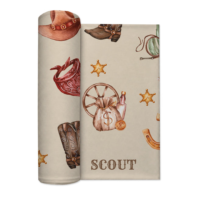 Partially rolled Wild West swaddle blanket with Scout lettering, cowboy boots, a wagon wheel and golden sheriff stars.
