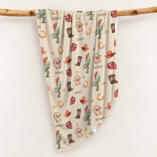 Personalized Wild West swaddle blanket draped over a wooden branch, showing its cream cowboy print and natural folds.