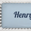 A light blue crib mattress fitted sheet with a white grid pattern and the partial name 'Henry' printed in dark navy script, shown inside a white wooden crib.