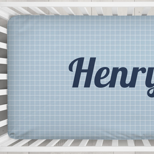 A light blue crib mattress fitted sheet with a white grid pattern and the partial name 'Henry' printed in dark navy script, shown inside a white wooden crib.
