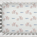A fitted crib sheet with a repeating pattern of pink pigs and the name 'Harlow' in blue, shown inside a white wooden crib.