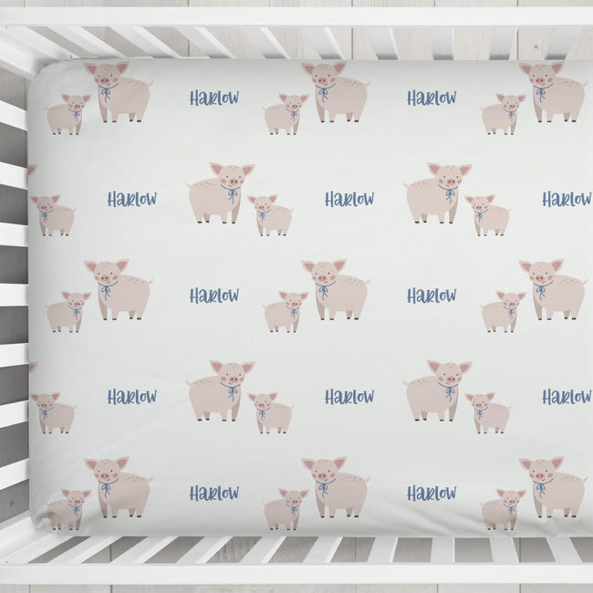 A fitted crib sheet with a repeating pattern of pink pigs and the name 'Harlow' in blue, shown inside a white wooden crib.