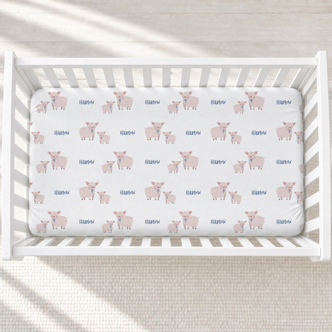 A white wooden crib with a fitted sheet featuring a repeating pattern of pink pigs and blue 'Harlow' text, photographed from above on a light textured floor.