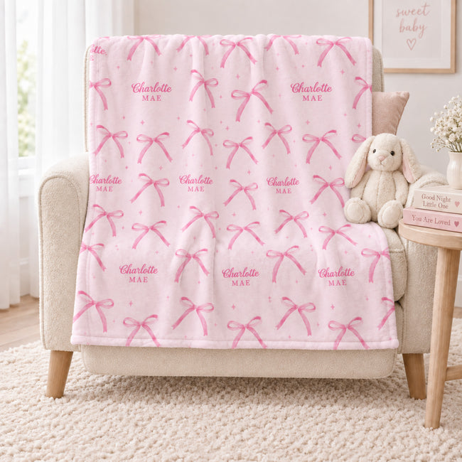 Personalized blush pink bow blanket with the repeated name Charlotte Mae draped over an ivory nursery chair beside a plush bunny.