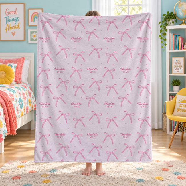 A child standing in a bedroom holding up a large light pink blanket or throw featuring a repeating pattern of pink bows and small pink stars, with the words 'Charlotte MAE' printed inside some bows.