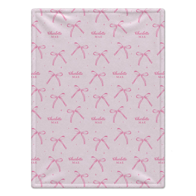 A baby blanket with a light pink background, featuring a repeating pattern of darker pink bows and small pink stars. Some names are personalized in the design, including 'Charlotte' and 'Mae'.