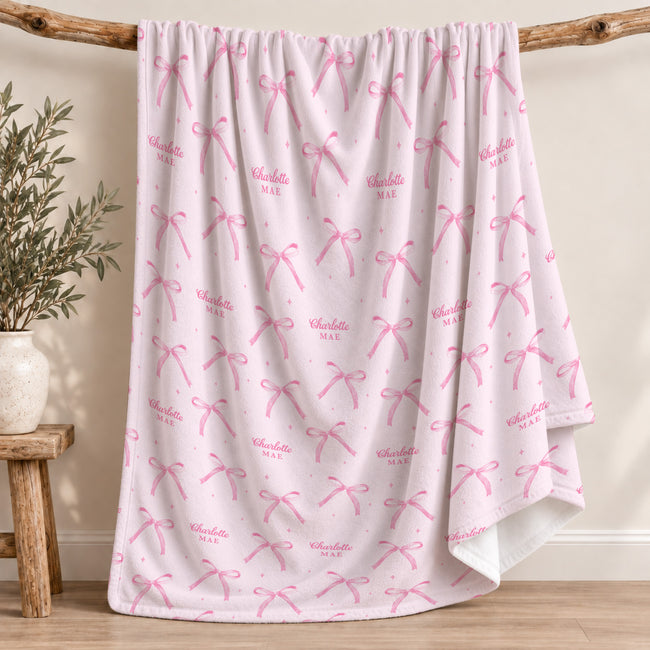 A light pink fleece blanket with a repeating pattern of pink bows and the names 'Charlotte MAE' in script, draped over a rustic wooden branch stand in a styled room setting.