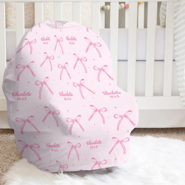 Blush pink baby car seat cover with watercolor bows, tiny sparkles, and the repeated name Charlotte Mae, shown in a bright nursery.