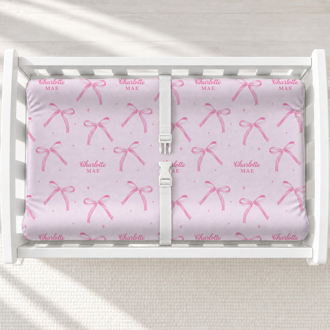 Personalized blush pink changing pad cover with watercolor bows, tiny sparkles, and the name Charlotte Mae on a white changing table.
