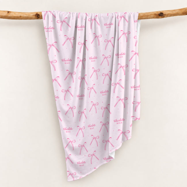 Blush pink personalized swaddle blanket with watercolor bows, tiny sparkles, and the name Charlotte Mae, draped over a wooden branch.