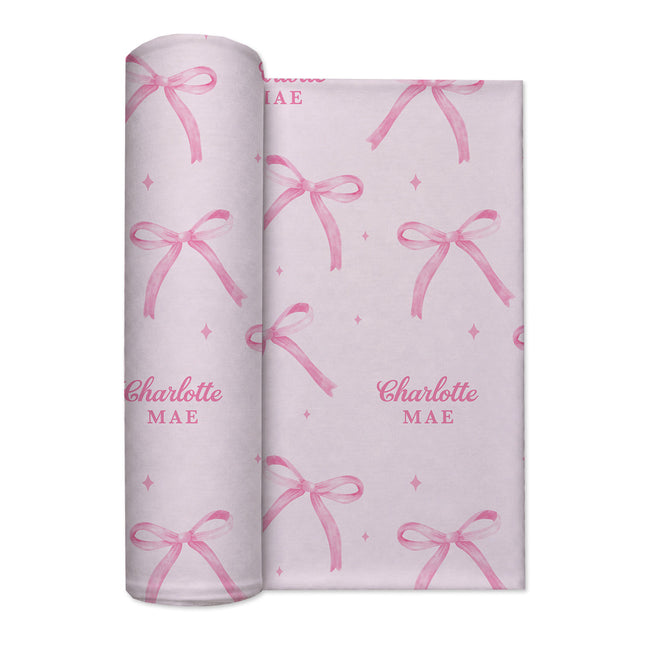 Rolled blush pink personalized swaddle blanket with watercolor bows, tiny sparkles, and the name Charlotte Mae.