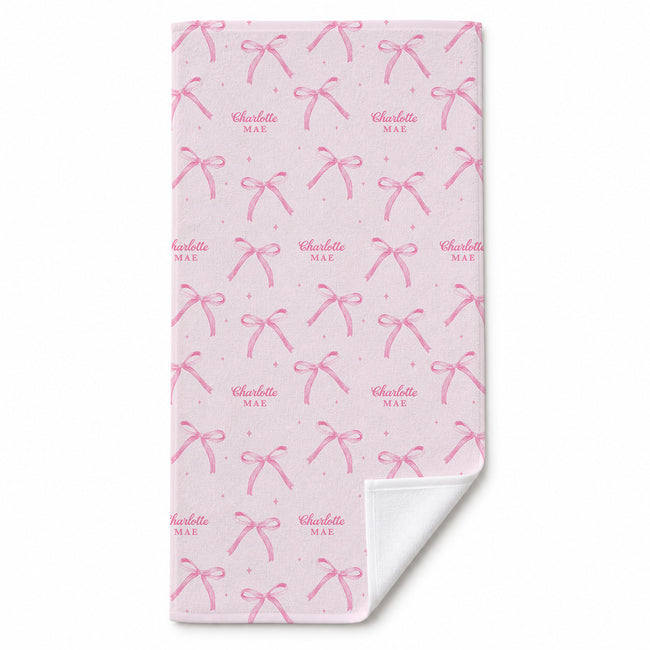 Blush pink beach towel with watercolor bows, tiny sparkles, and the repeated name Charlotte Mae, with a folded corner showing the white reverse.