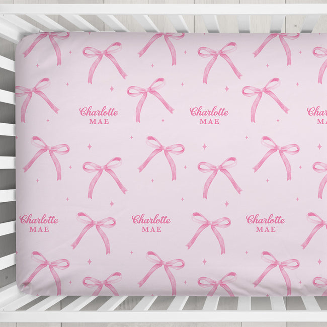 Personalized blush pink crib sheet with watercolor bows, tiny sparkles, and the repeated name Charlotte Mae, shown inside a white crib.