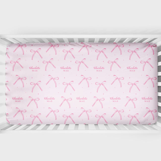 Personalized blush pink crib sheet with watercolor bows, tiny sparkles, and the repeated name Charlotte Mae, shown inside a white crib.
