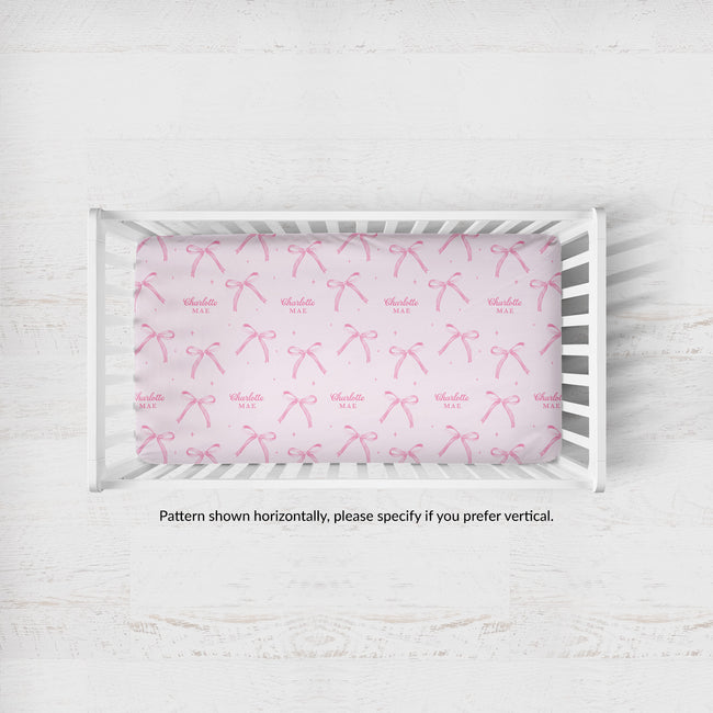 Personalized blush pink crib sheet with watercolor bows, tiny sparkles, and the repeated name Charlotte Mae, shown inside a white crib.