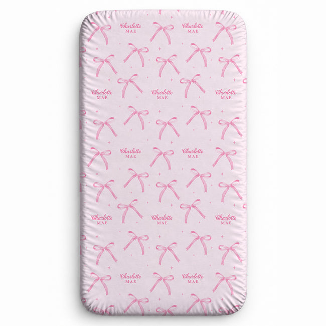 Blush pink twin fitted sheet with watercolor bows, tiny sparkles, and the name Charlotte Mae repeated in pink script and uppercase lettering.