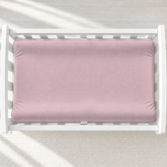A dusty pink changing pad cover inside a white wooden baby changing table, shown from a top-down view on a light textured floor with soft diagonal shadows.