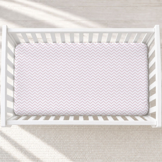 A top-down view of a white wooden crib with a fitted sheet featuring a light pink and white chevron pattern, set against a neutral cream background with soft diagonal shadows.