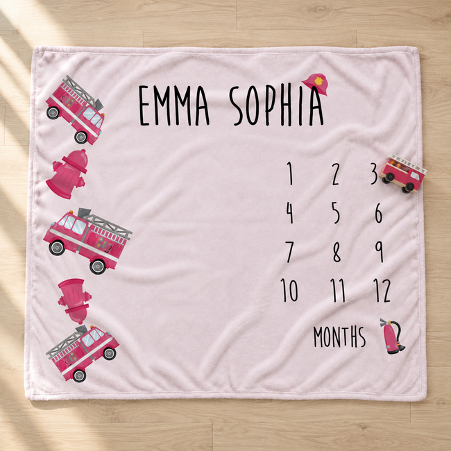 A personalized baby blanket laid flat on a light wood floor. The blanket is pale pink with black text reading 'EMMA SOPHIA' and a 12-month countdown grid labeled 'MONTHS'.