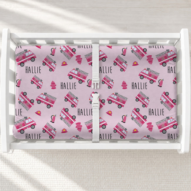 A white changing table with a personalized changing pad cover featuring a pink and gray fire truck pattern with the name 'HALLIE' repeated. 