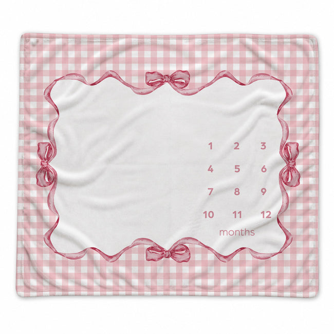 A baby milestone blanket with a pink and white gingham border, a scalloped pink frame, ribbon-bow details, and a central white area printed with numbers 1 through 12 and the word 'months' for tracking birth months.