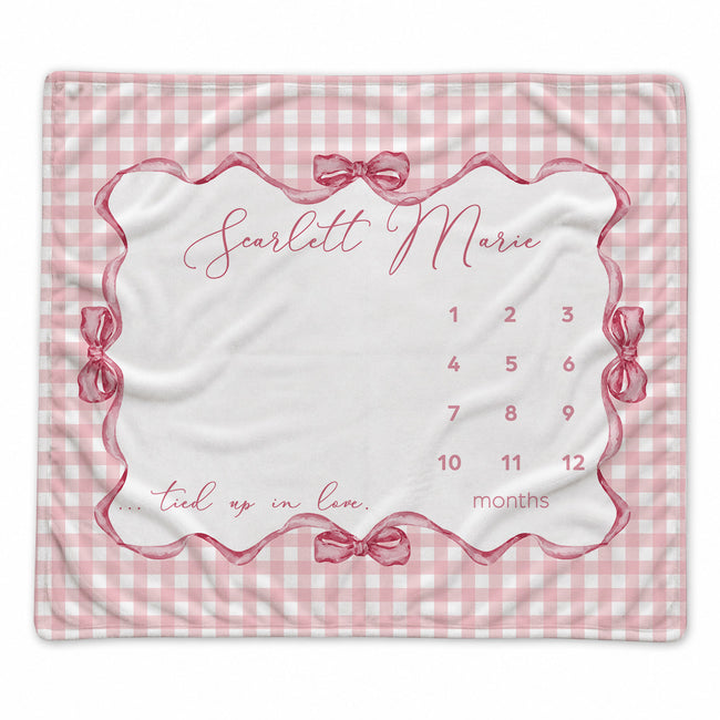 A baby milestone blanket with a pink and white gingham border, featuring a central white panel with a scalloped pink frame, bows, the name 'Scarlett Marie' in script, and a numbered 1-12 timeline for tracking months.