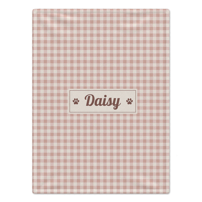 A rectangular pink and white gingham dog blanket with a central cream label that reads 'Daisy' in brown script, flanked by two small paw prints, shown on a plain white background.