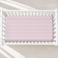 A top-down view of a white wooden crib with a fitted sheet featuring horizontal pink and white stripes, set against a light beige textured rug with soft window shadow patterns.