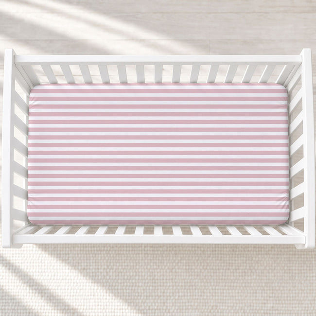 A top-down view of a white wooden crib with a fitted sheet featuring horizontal pink and white stripes, set against a light beige textured rug with soft window shadow patterns.