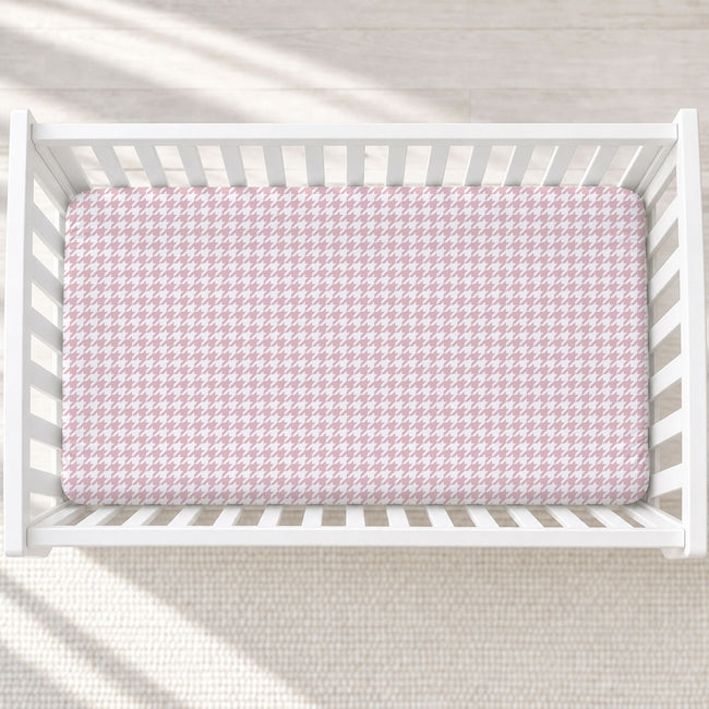 A top-down view of a white wooden crib containing a mattress fitted with a light pink and white houndstooth pattern sheet, set against a neutral cream textured background.