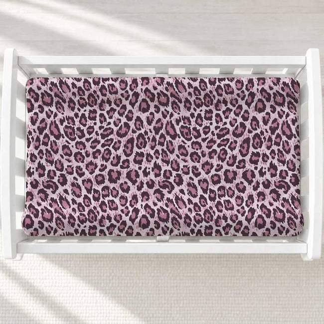 A changing pad cover with a pink and purple leopard print pattern displayed inside a white wooden baby crib, viewed from a top-down perspective.